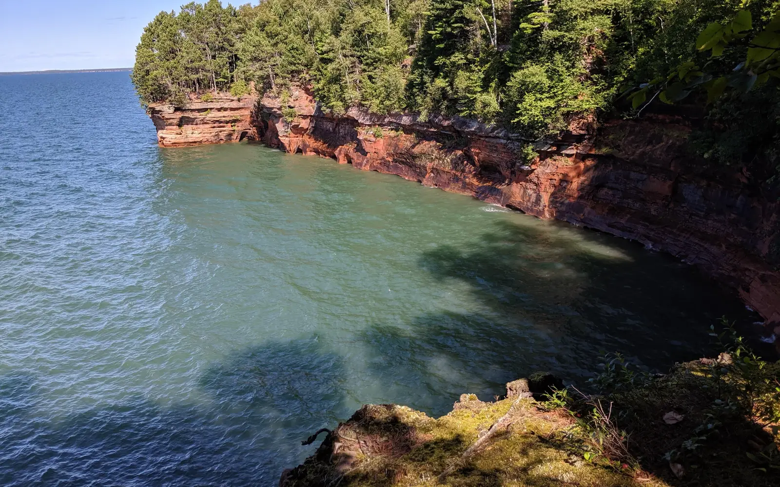 Tree-covered red cliffs meet greenish blue water, in a bowl shape.
