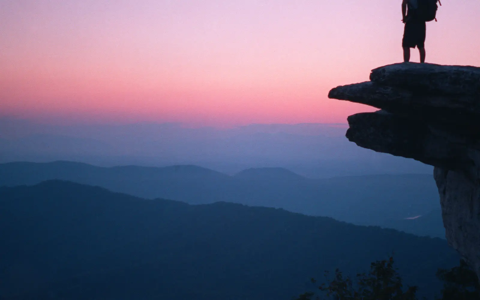 Silhouette of a man with backpack standing on McAfee Knob at sunset with mountains in the distance.