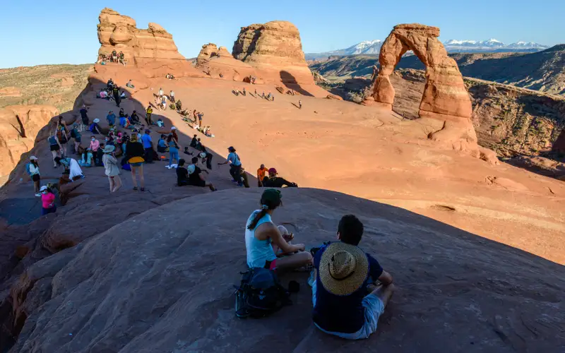 A crowd of people sit and watch the sunset at delicate arch.