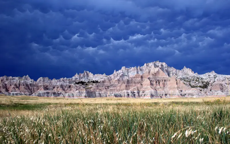 Layered badlands formations behind fields of green grass under cloudy and billowing clouds.