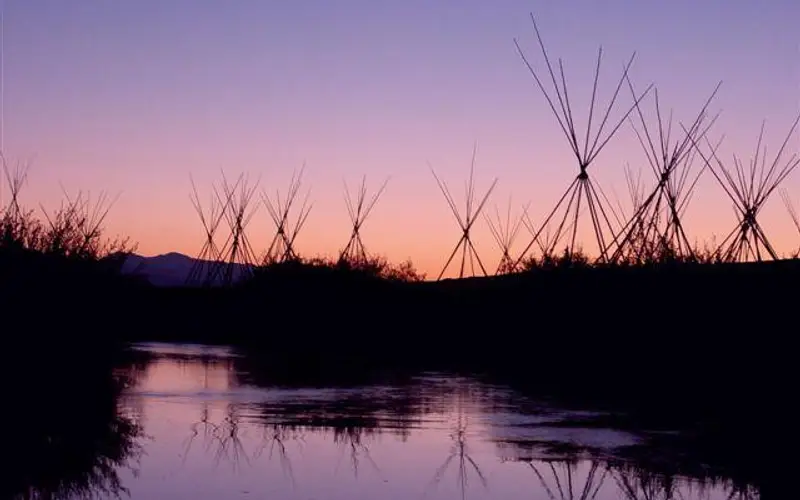 Multiple tepee poles and a river are silhouetted against a dawn sky.