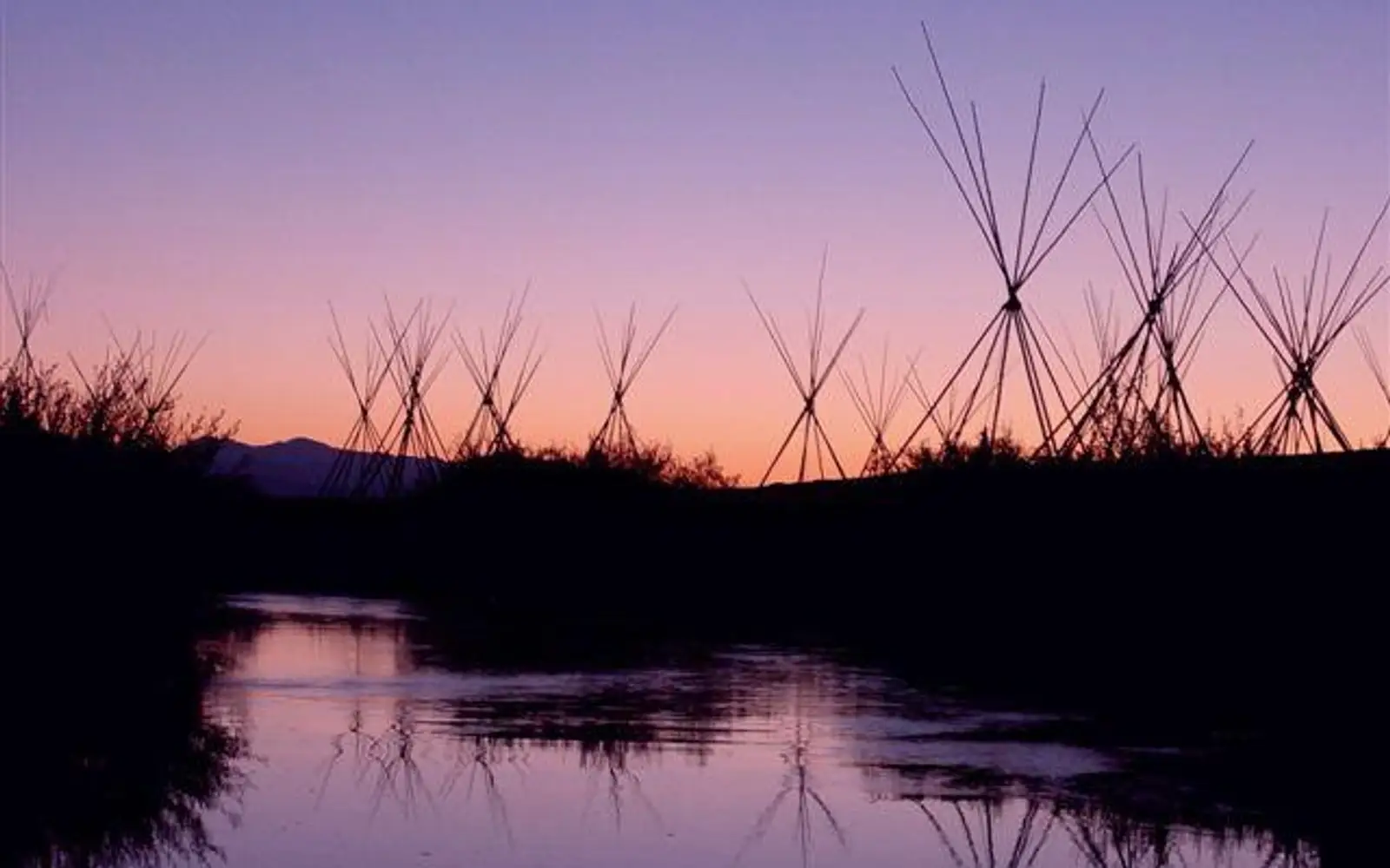 Multiple tepee poles and a river are silhouetted against a dawn sky.