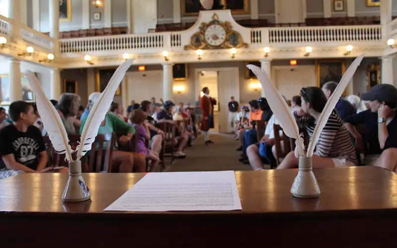 A desk with paper and two sets of quills frame a person in colonial wear standing in the aisle.