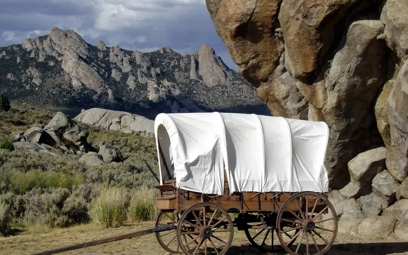 A white canvas wagon sits in front of a large rock buttress with mountains in the distance.