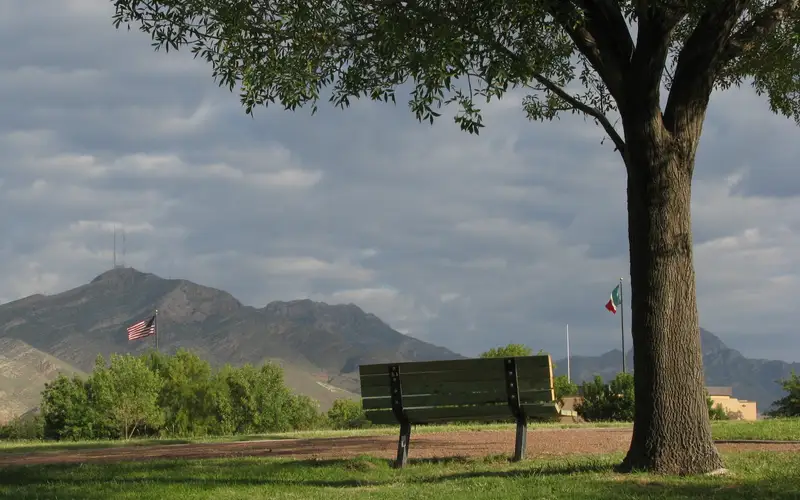 a bench next to a tree with U.S. and Mexican flags flying against a mountain background
