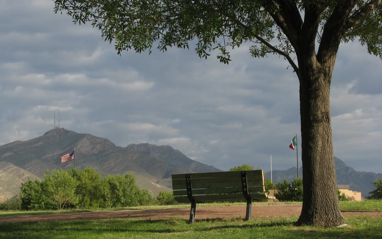 a bench next to a tree with U.S. and Mexican flags flying against a mountain background
