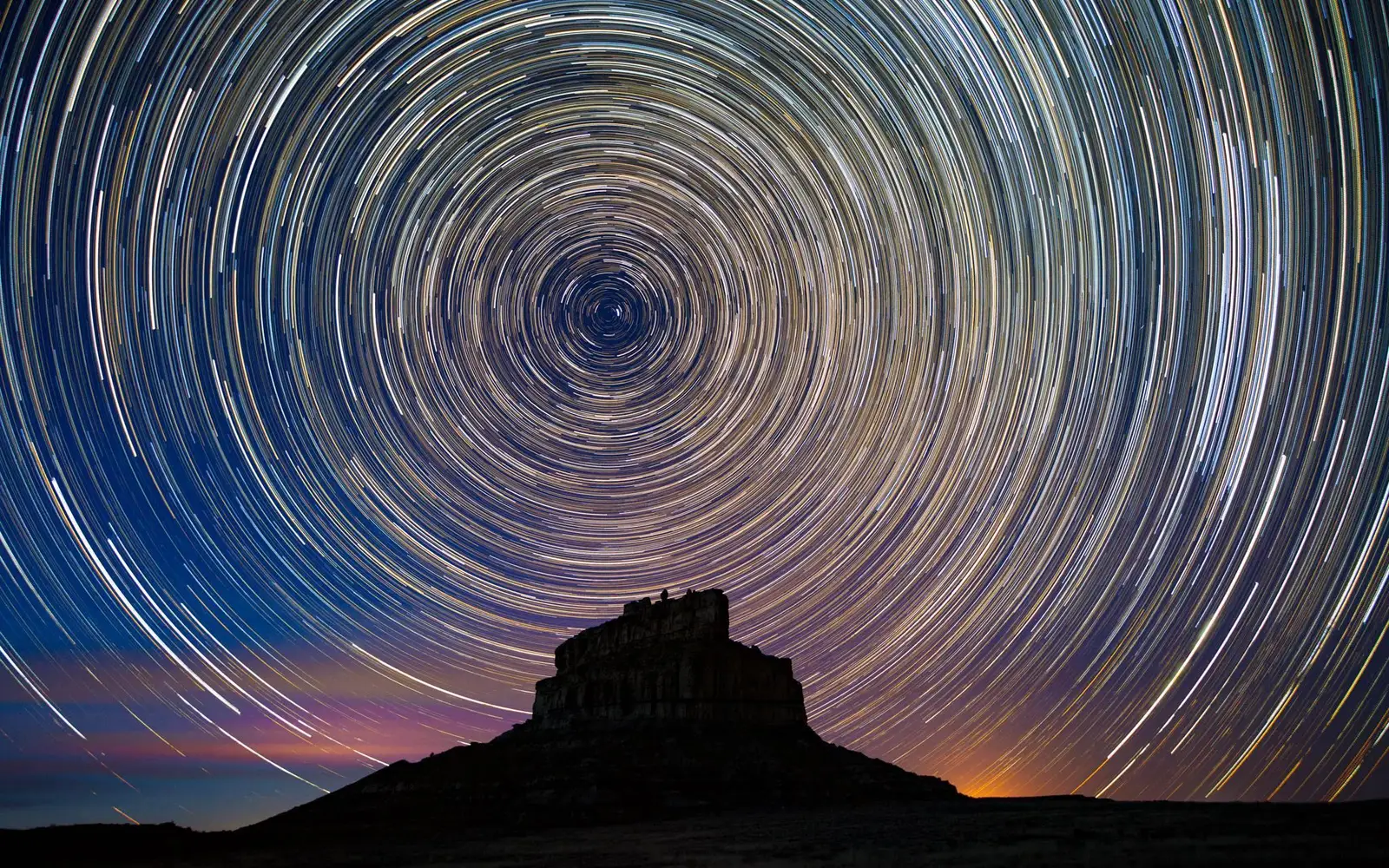 Fajada Butte's silhoutte shown under a timelapse image of stars appearing in a vibrant circle above.