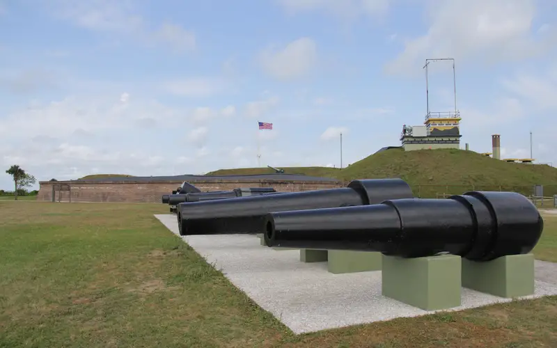 A row of cannon with Fort Moultrie in the background. A US flag is flying in the fort.