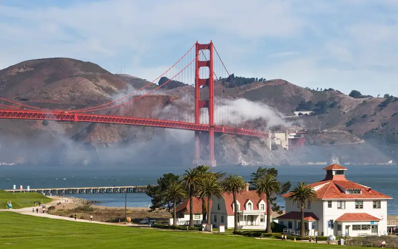 White buildings with red roofs at Crissy Field with blue bay and Golden Gate Bridge and fog behind.