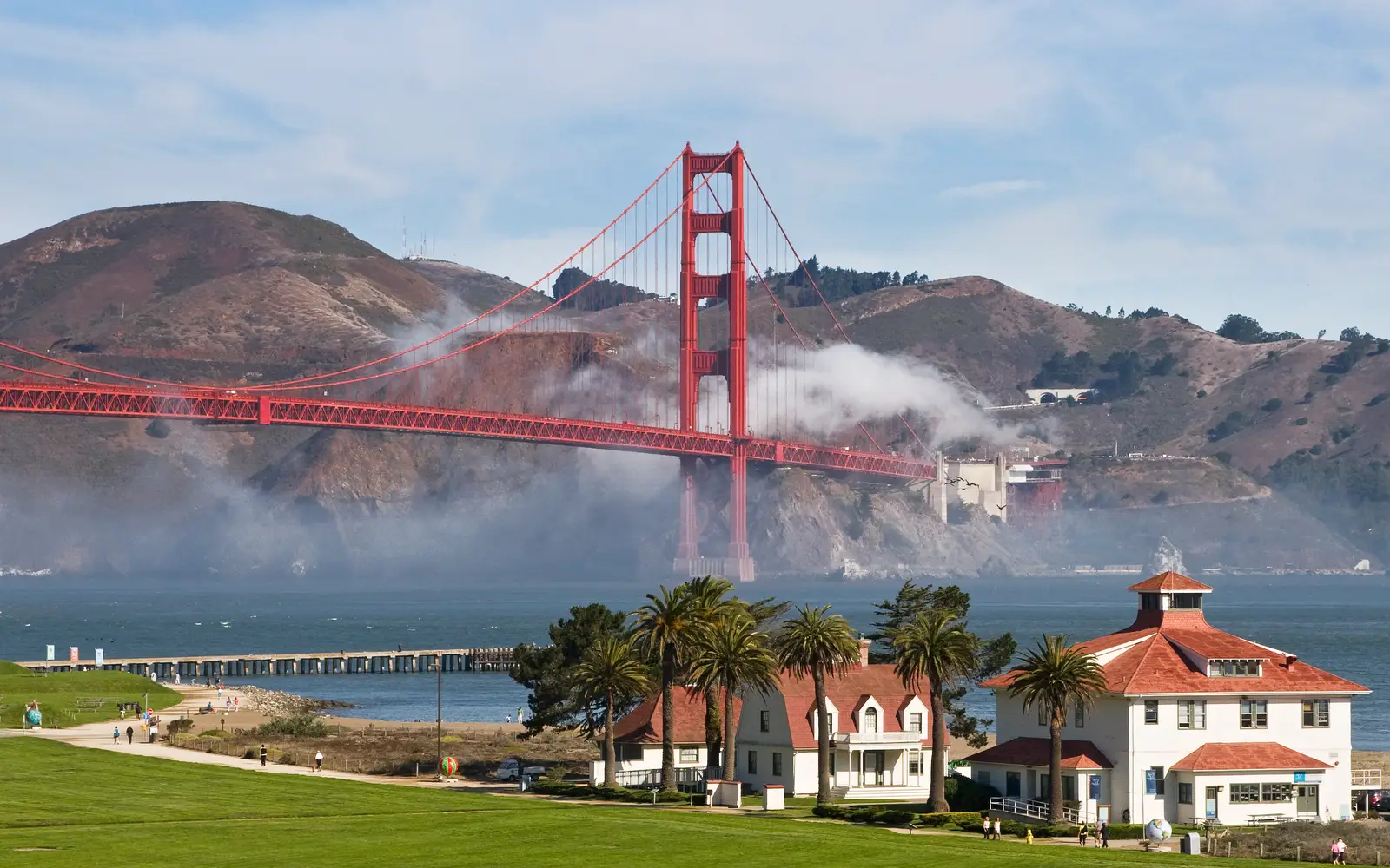 White buildings with red roofs at Crissy Field with blue bay and Golden Gate Bridge and fog behind.