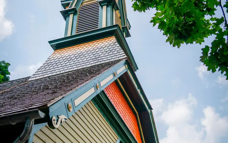 A tall, green and orange painted church steeple, against a blue sky, with a tree in the foreground.