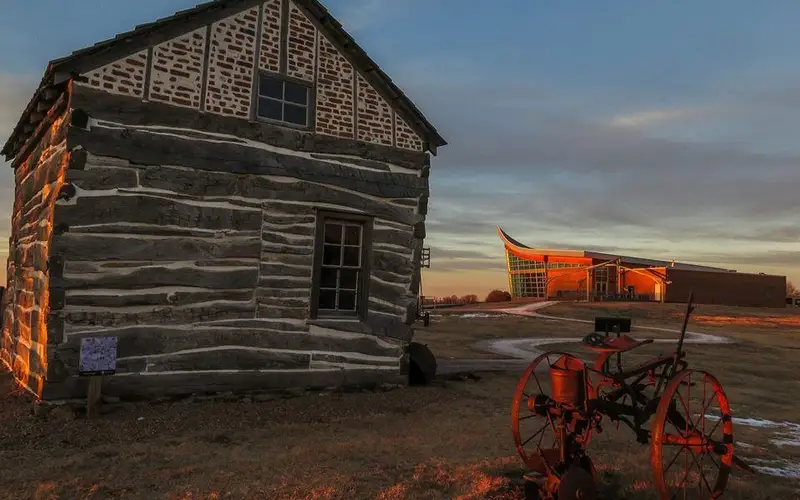 A historic cabin stands next to a concrete path that leads to a plow shaped visitor center