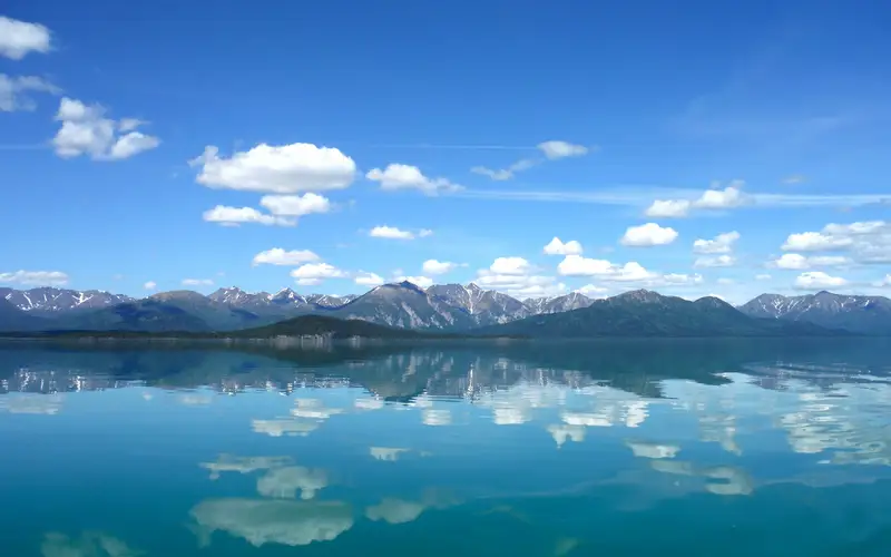 Photo of blue sky with fluffy white clouds reflect in calm lake with mountains in the background.