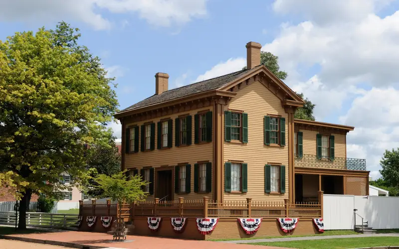 2 story tan house with green shutters. Red, white, and blue striped bunting draped below its fence
