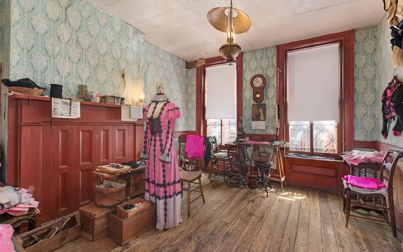 A room with wooden floors, blue wall paper, two windows, some furniture and a dress hanging.