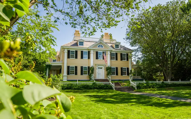 Yellow three-story mansion with symmetrical facade. Steps and large lawn in foreground. Framed by br