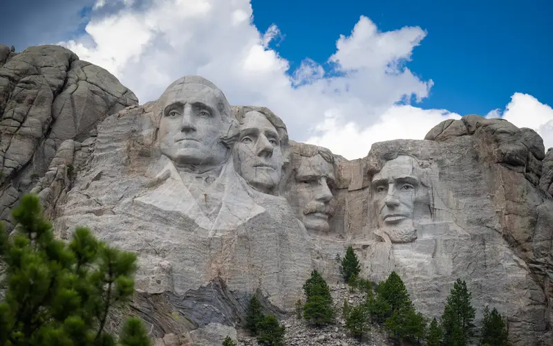Photo of Mount Rushmore under a bright blue sky with puffy clouds floating over.