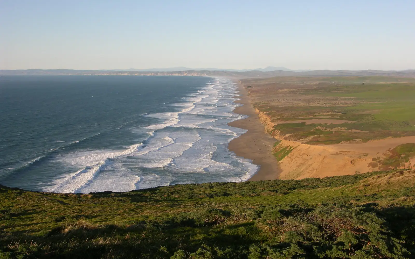 Waves wash in from the left onto a long, straight stretch of undeveloped, low-lying coastline.