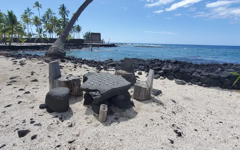 A stone papamū (Kōnane playing surface) with Hale o Keawe and Hōnaunau Bay in the background.