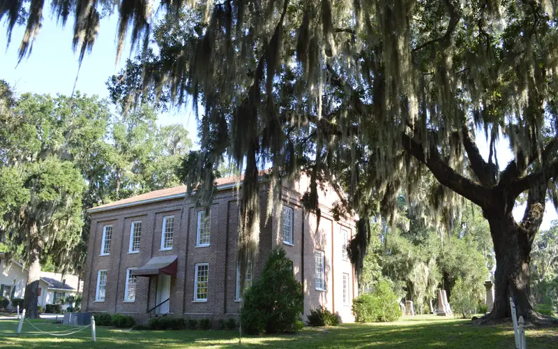 A two story brick building flanked by large live oak trees