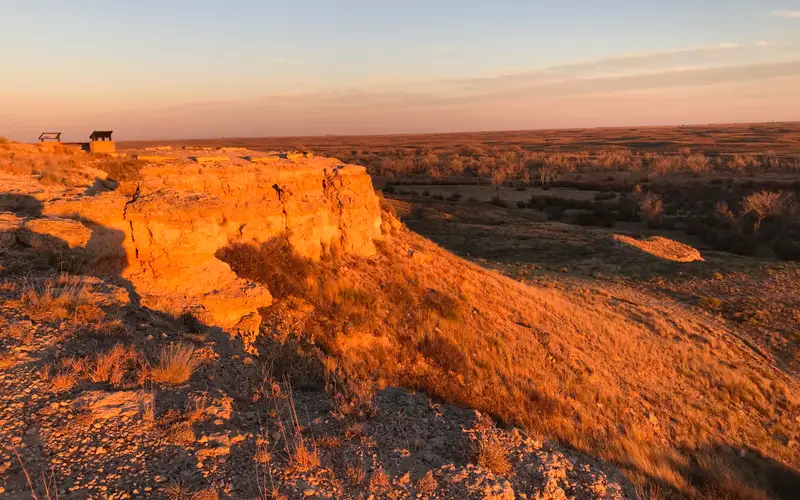 Warm light from the setting sun gives an orange glow to a rock bluff overlooking a grassland.