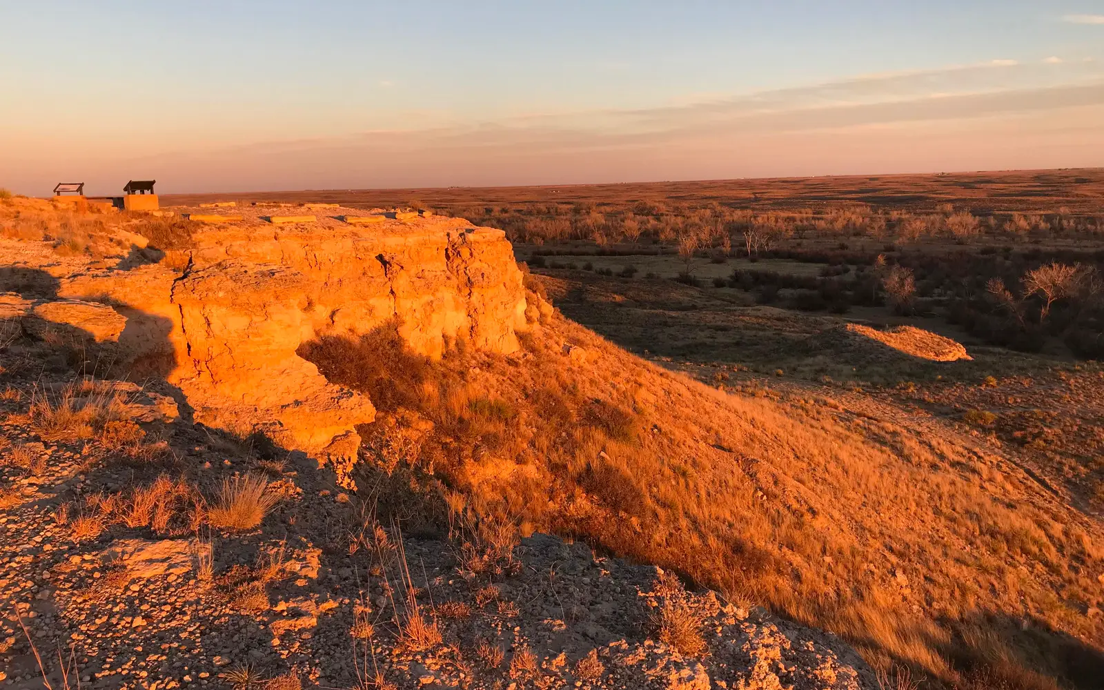 Warm light from the setting sun gives an orange glow to a rock bluff overlooking a grassland.