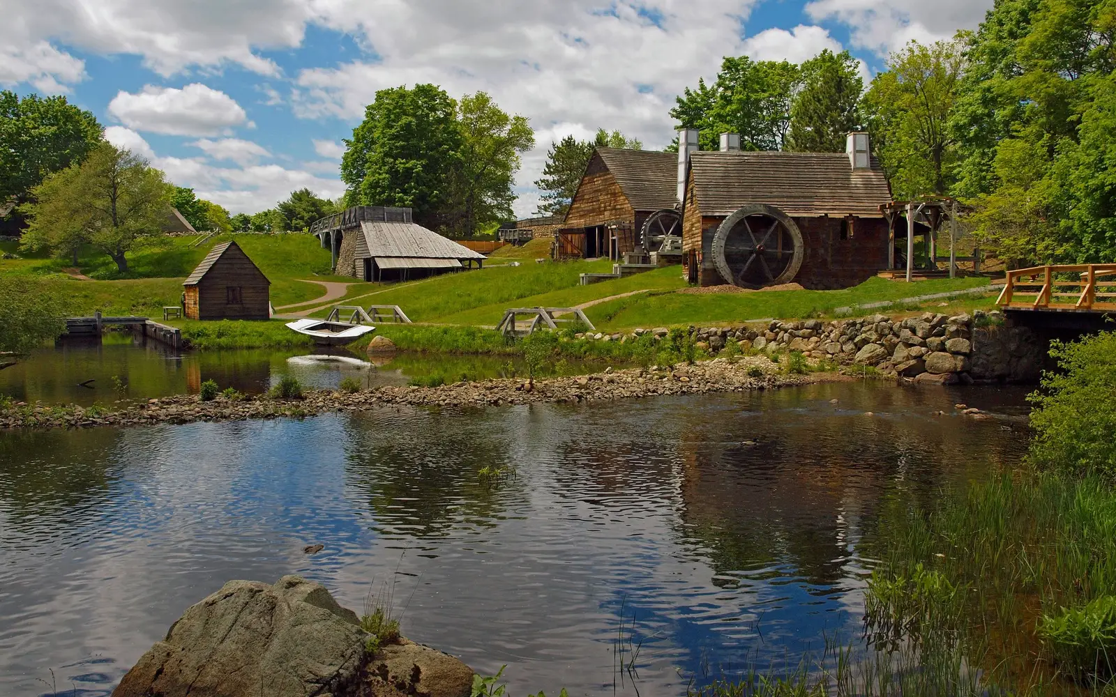 Several wooden structures amid green fields and trees under partly cloudy sky and beside river.