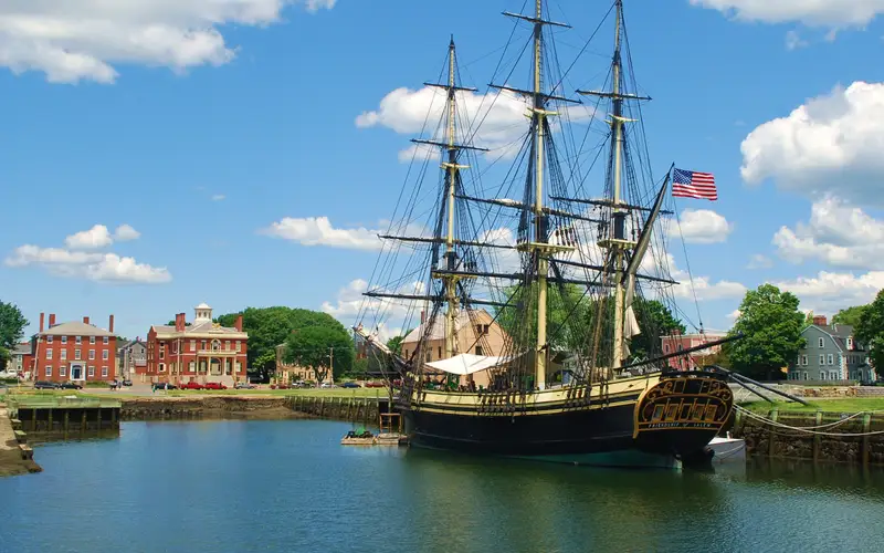 A three mast tall ship on the water under a blue sky with red brick buildings on the shore.