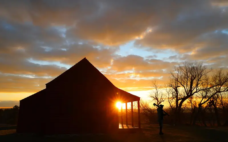 A man wearing 18th-century clothing carrying a musket and a farmhouse are silhouetted by sunrise.
