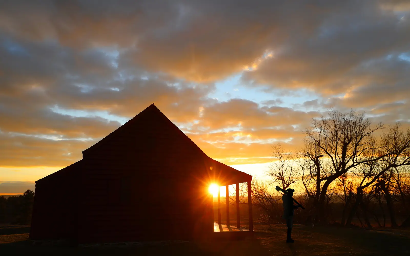 A man wearing 18th-century clothing carrying a musket and a farmhouse are silhouetted by sunrise.