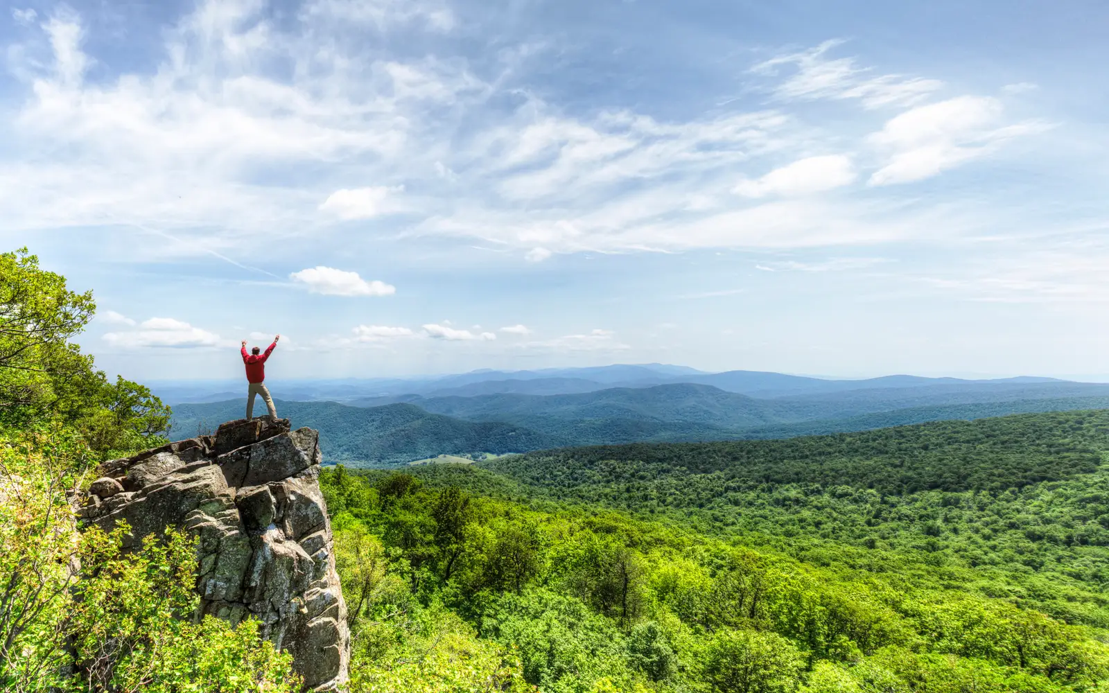 A man stands on a rocky outcrop overlooking the receding mountains.
