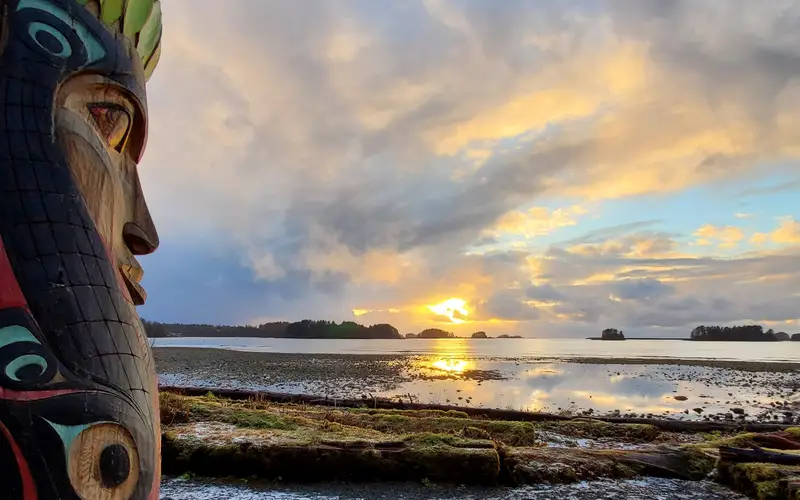 A totem pole in the foreground with a sunset over water in the background.