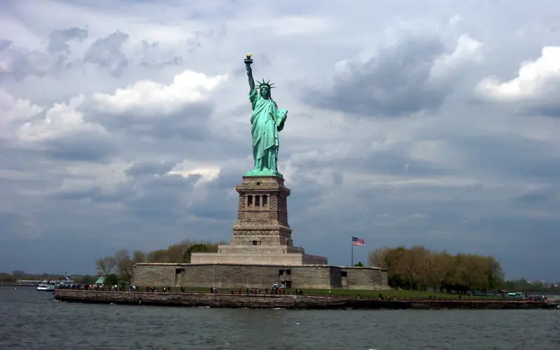 Patina green statue atop her pedestal on Liberty Island; grey cloudy sky in the distance