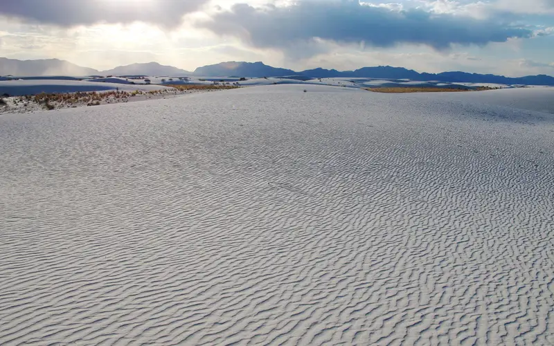 White dunes in foreground with sun setting behind mountain.