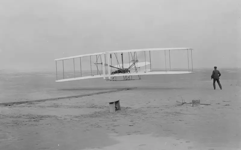 Orville takes to the air in the flyer for the first time as Wilbur assists.