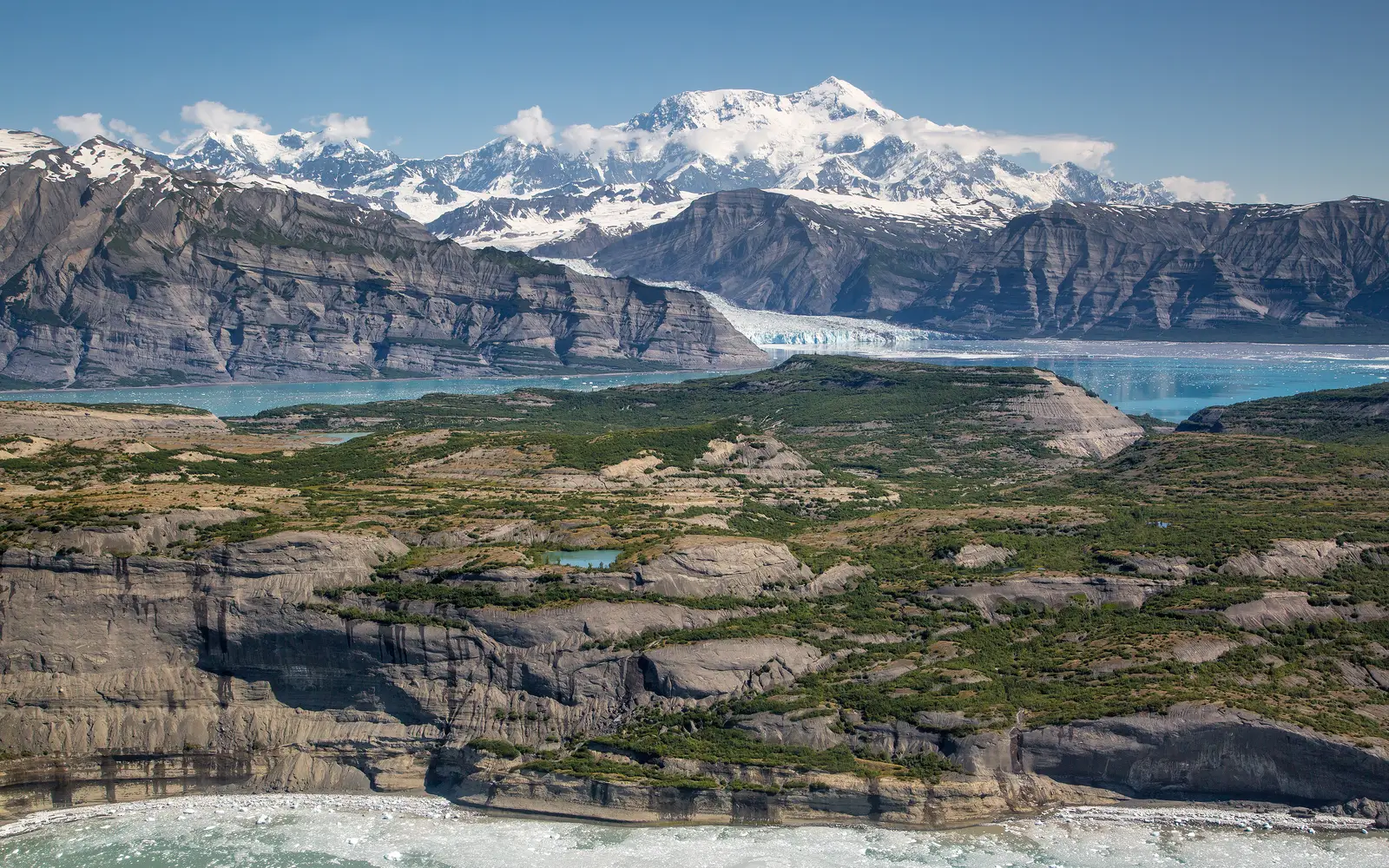 Glaciers loom over the ocean with large snowy mountains rising into blue skies