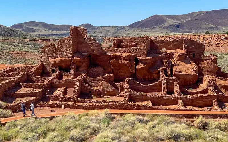 a large stone pueblo in a desert beneath mostly sunny skies with mountains in background